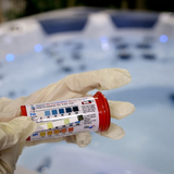 A gloved hand holding a Poppits Peroxsil test strip against the detailed bottle instructions, showing the peroxide ppm, alkalinity, and pH color scales over a lit spa.