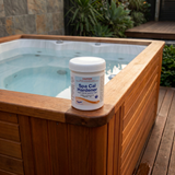 A 500g container of Poppits Spa Cal Hardener sitting on the wooden corner ledge of an outdoor hot tub. The label shows it raises calcium hardness levels to protect the spa, with clear water visible in the background deck setting.