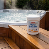 A 1kg tub of Poppits Water Balance sitting on a wooden step next to a steaming outdoor hot tub, used to raise pH and total alkalinity in chlorine-free spas.