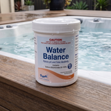 A 1kg container of Poppits Water Balance sitting on a wooden deck next to a bubbling spa. The label identifies it as Step 2, containing 99-100% Sodium Bicarbonate, used to raise pH and Total Alkalinity in hot tubs and pools.