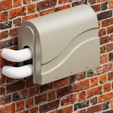 Angled side view of a beige Protec spa bath pump cover mounted against a red brick exterior wall, protecting two white PVC elbow pipe connections.