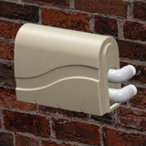 In-situ lifestyle image showing a beige Protec spa bath pump cover mounted against a red brick exterior wall over two white PVC elbow pipes.