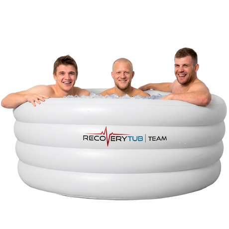 Three smiling male athletes sitting in a large white inflatable Recovery Tub Team ice bath filled with ice cubes for post-workout muscle recovery.