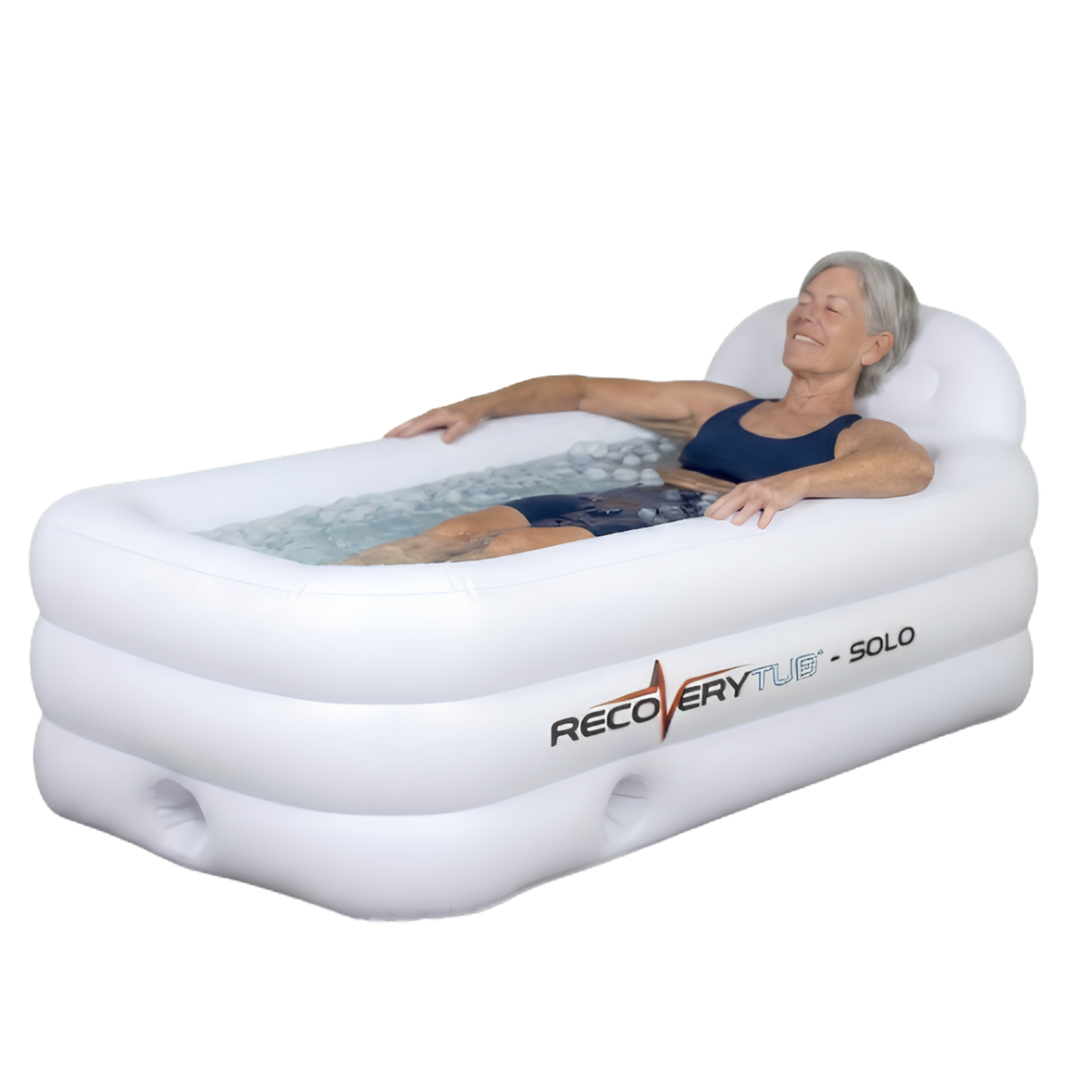 A senior female athlete relaxing with her eyes closed inside a white rectangular RecoveryTub Solo inflatable ice bath filled with ice water, resting her head against the built-in headrest, isolated on a white background