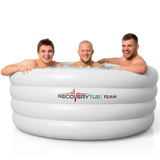 Three smiling male athletes sitting in a large white inflatable RecoveryTub Team ice bath filled with ice cubes and water for post-workout athletic recovery, isolated on a white background.