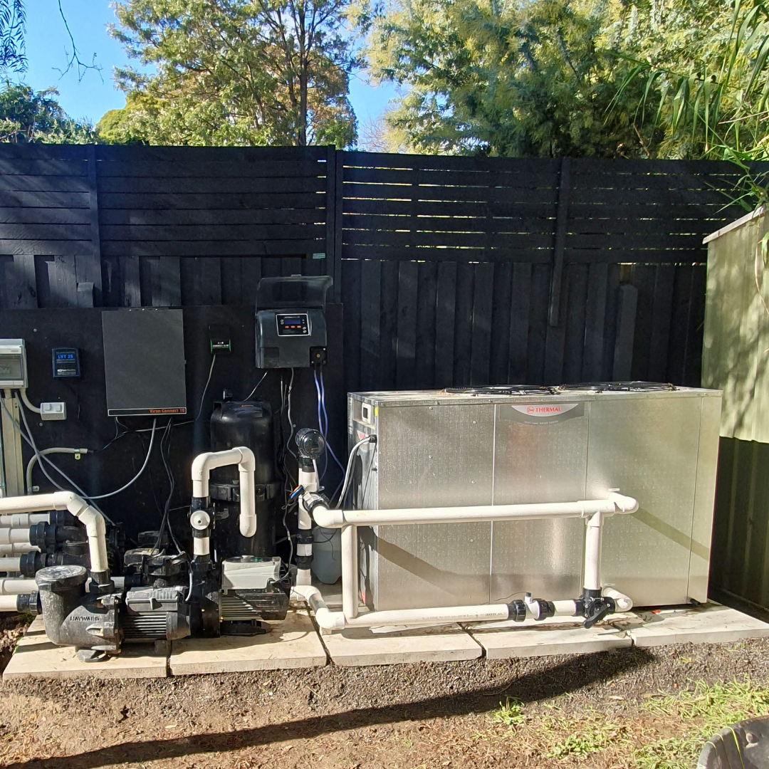 Outdoor backyard pool equipment installation featuring a commercial-grade Rheem Thermal top-discharge pool heat pump on a paved concrete pad alongside filtration pumps, PVC plumbing, and wall-mounted control panels against a black wooden fence.