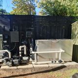 Outdoor backyard pool equipment installation featuring a commercial-grade Rheem Thermal top-discharge pool heat pump on a paved concrete pad alongside filtration pumps, PVC plumbing, and wall-mounted control panels against a black wooden fence.