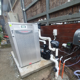 Residential outdoor installation of a Rheem Thermal pool heat pump set on a concrete slab, showcasing white PVC plumbing pipes, water circulation pump, electrical connections, and wall-mounted controllers against a wooden retaining fence.
