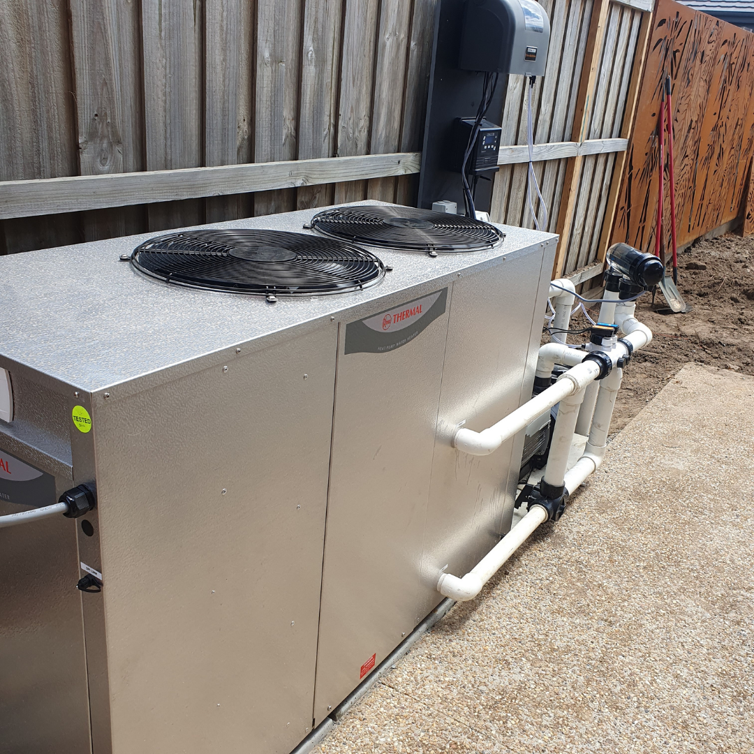 Outdoor real-world installation of a Rheem Thermal top-discharge pool heat pump unit set on a concrete pad alongside a timber fence, complete with PVC pipe connections and wall-mounted pool control equipment.