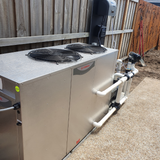 Outdoor real-world installation of a Rheem Thermal top-discharge pool heat pump unit set on a concrete pad alongside a timber fence, complete with PVC pipe connections and wall-mounted pool control equipment.