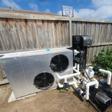 Outdoor backyard installation of a Rheem Thermal side-discharge pool heat pump featuring dual fan grilles, positioned along a wooden boundary fence with PVC plumbing pipes, pumps, and wall-mounted controller equipment.
