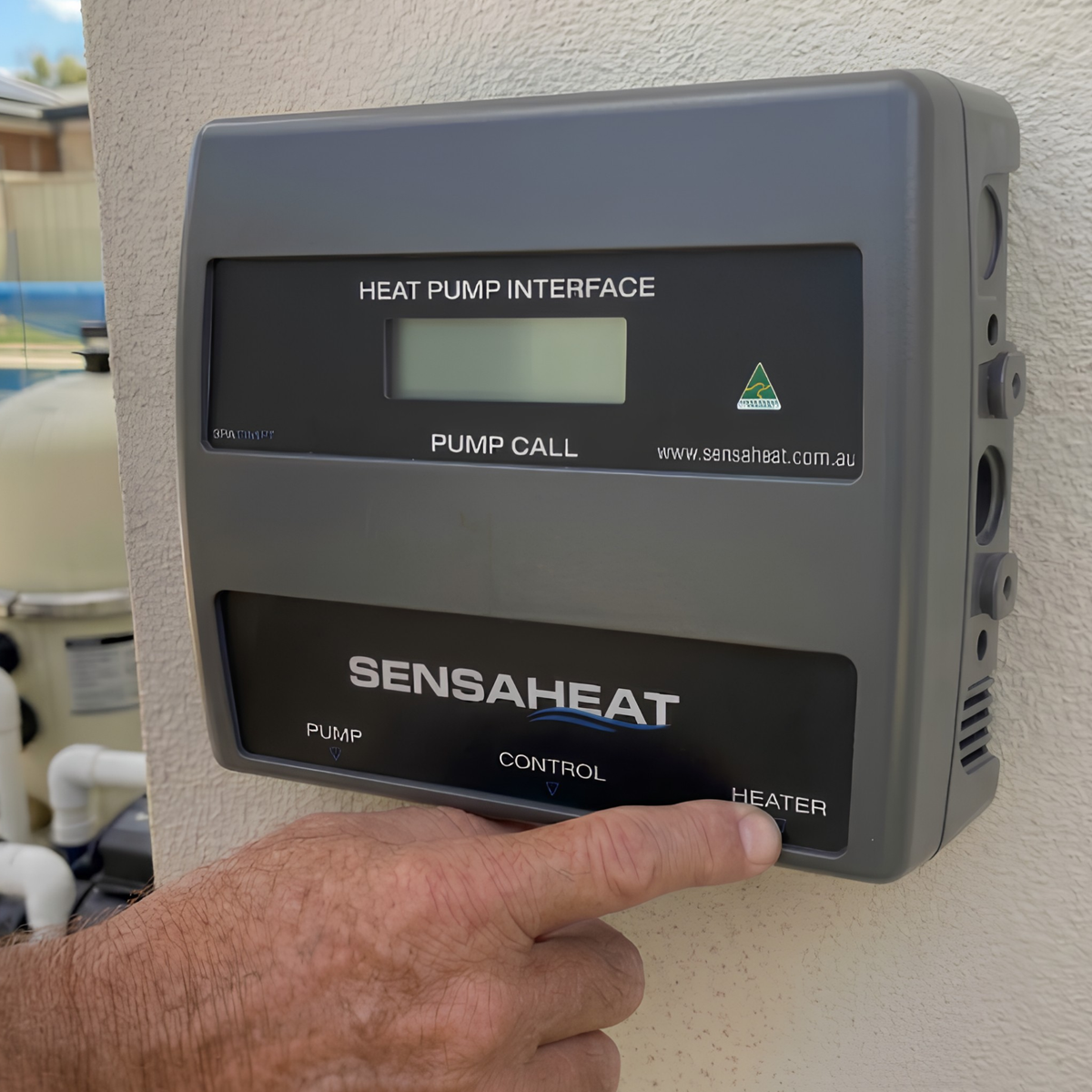 Close-up of a wall-mounted Sensaheat Heat Pump Interface pool automation controller outdoors next to a pool filter, with a user's finger pressing the lower front control panel.