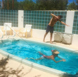 A woman wearing a visor training in a compact outdoor plunge pool using a Swim Tether system, attached by a black resistance cord to a pole on the deck while a man stands on the poolside watching, with a blue glass block wall in the background.