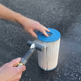 Action photo of a person using a grey Spa AquaComb tool to spray water and comb between the pleats of a small white spa cartridge filter, set against an asphalt ground background.
