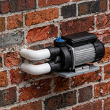 In-situ lifestyle image showing an uncovered spa bath pump mounted on a metal wall bracket against a red brick exterior wall with two white PVC elbow pipe connections.