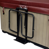 A close-up view of a black metal gas spring-assisted spa cover lifter mechanism mounted to the tan wood-paneled side of a hot tub with a closed brown leather cover, isolated on a white background.