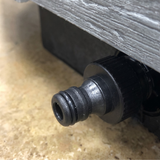Close up angled view of a black plastic spa drain tap connector adapter screwed into the lower edge of a grey wood grain hot tub cabinet panel