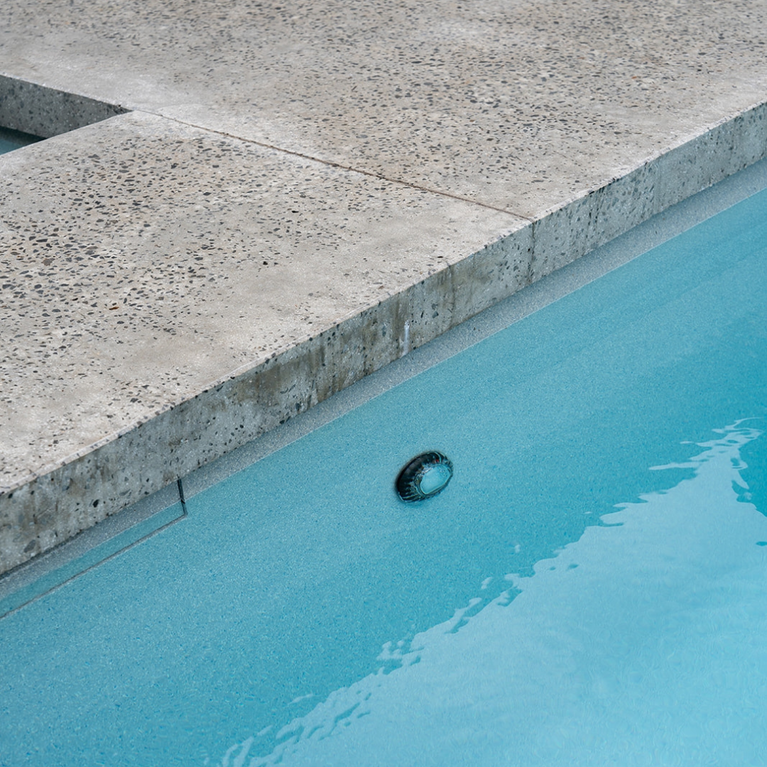 Close-up daytime lifestyle image of a single Spa Electrics EMF Multi Plus LED pool light installed underwater along the interior wall of a swimming pool with exposed aggregate concrete coping.
