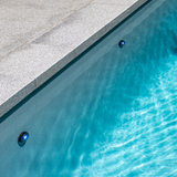 In-situ lifestyle image showing two Spa Electrics EMF Multi Plus LED pool lights emitting blue illumination underwater along the wall of a concrete pool with grey stone coping.