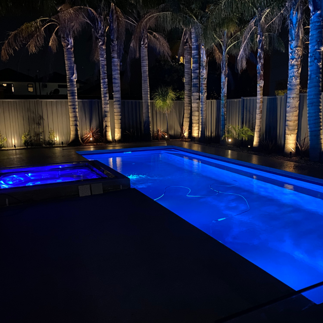 Nighttime lifestyle view of a residential swimming pool and adjoining spa brightly illuminated in vibrant blue by Spa Electrics EMF Multi Plus LED lights with palm trees in the background.