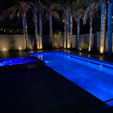 Nighttime lifestyle view of a residential swimming pool and adjoining spa brightly illuminated in vibrant blue by Spa Electrics EMF Multi Plus LED lights with palm trees in the background.