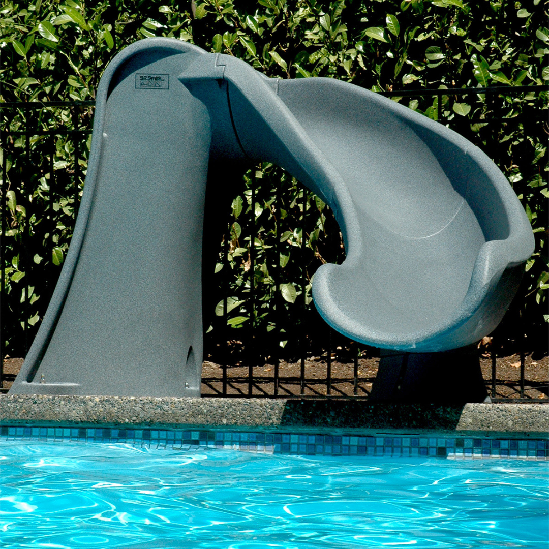 A poolside lifestyle installation shot of an S.R. Smith Cyclone deck-mounted pool slide in gray granite color. The slide curves sharply to the right into the blue shimmering pool water, set against a concrete pool coping edge, black metal security fence, and lush green hedge bushes.