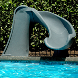 A poolside lifestyle installation shot of an S.R. Smith Cyclone deck-mounted pool slide in gray granite color. The slide curves sharply to the right into the blue shimmering pool water, set against a concrete pool coping edge, black metal security fence, and lush green hedge bushes.