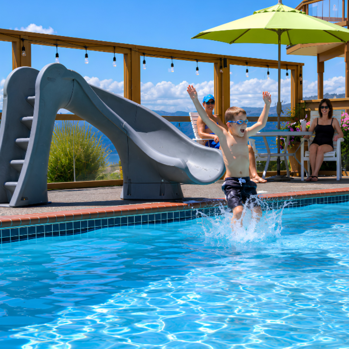 A vibrant backyard lifestyle shot of a young boy splashing into a blue inground swimming pool after sliding down a gray granite S.R. Smith Cyclone deck-mounted pool slide. In the background, two adults watch from patio chairs underneath a bright green umbrella on a sunny pool deck.