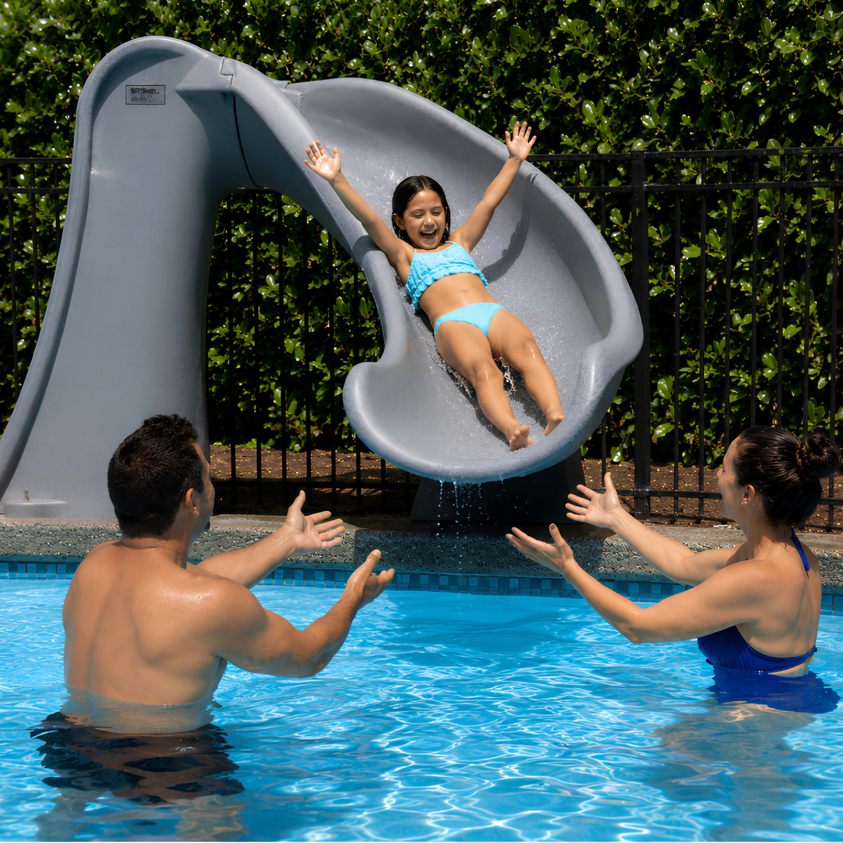 A joyful family lifestyle shot of a young girl in a blue bikini sliding down a gray granite S.R. Smith Cyclone deck-mounted pool slide with her arms raised. Her parents stand in the bright blue swimming pool water with outstretched arms to catch her, set against a black safety fence and a lush green hedge.