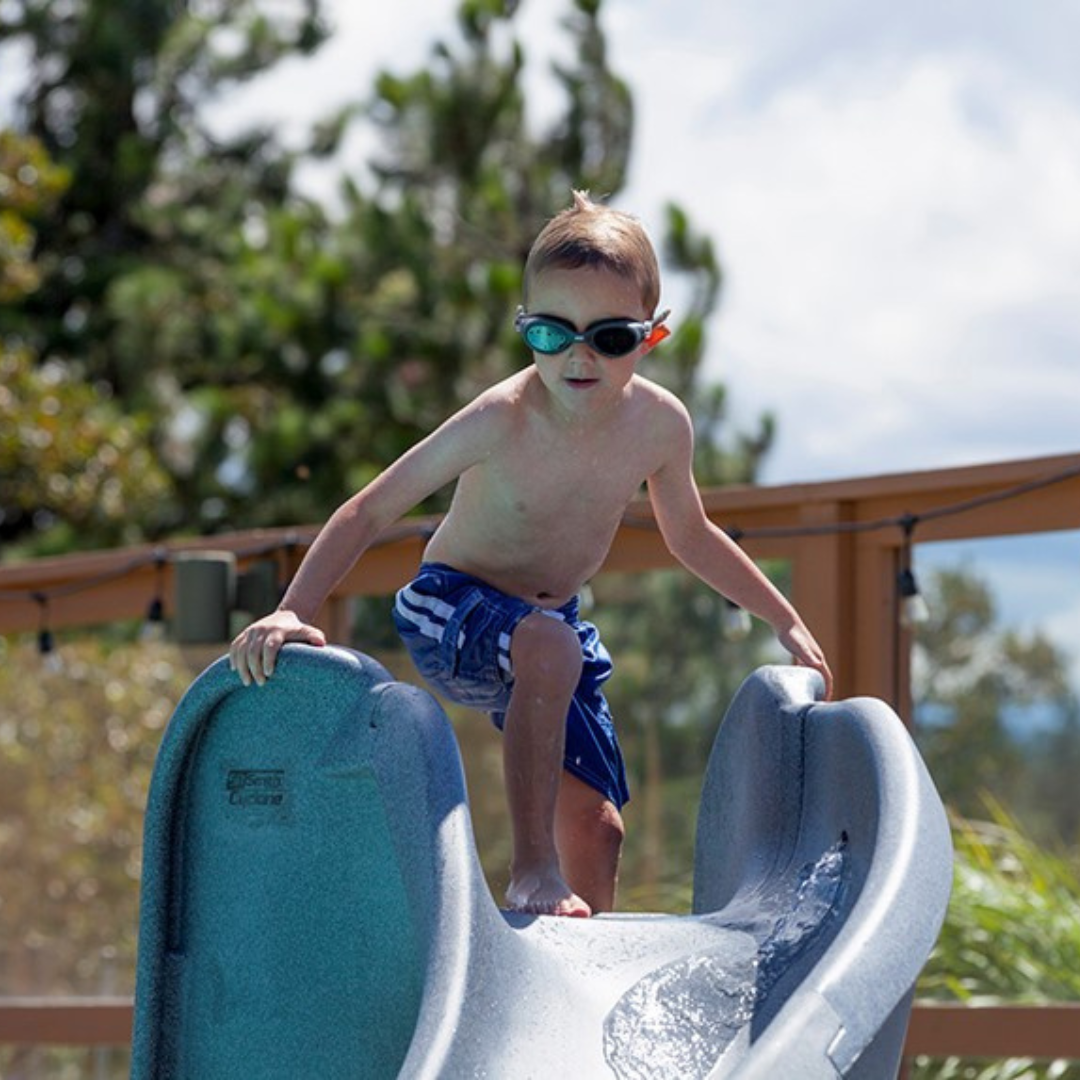 A close-up lifestyle shot of a young boy wearing swim goggles preparing to slide down a gray granite S.R. Smith Cyclone deck-mounted pool slide, with the molded manufacturer logo clearly visible on the side flange against a sunny backyard background.
