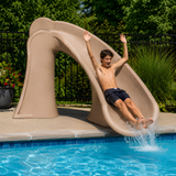An energetic lifestyle shot of a teenage boy in black swim shorts sliding down a tan sandstone granite S.R. Smith Cyclone deck-mounted pool slide with his arms raised. The water slide curves right over a blue tiled inground swimming pool, bordered by a concrete patio, a black security fence, and lush green bushes.