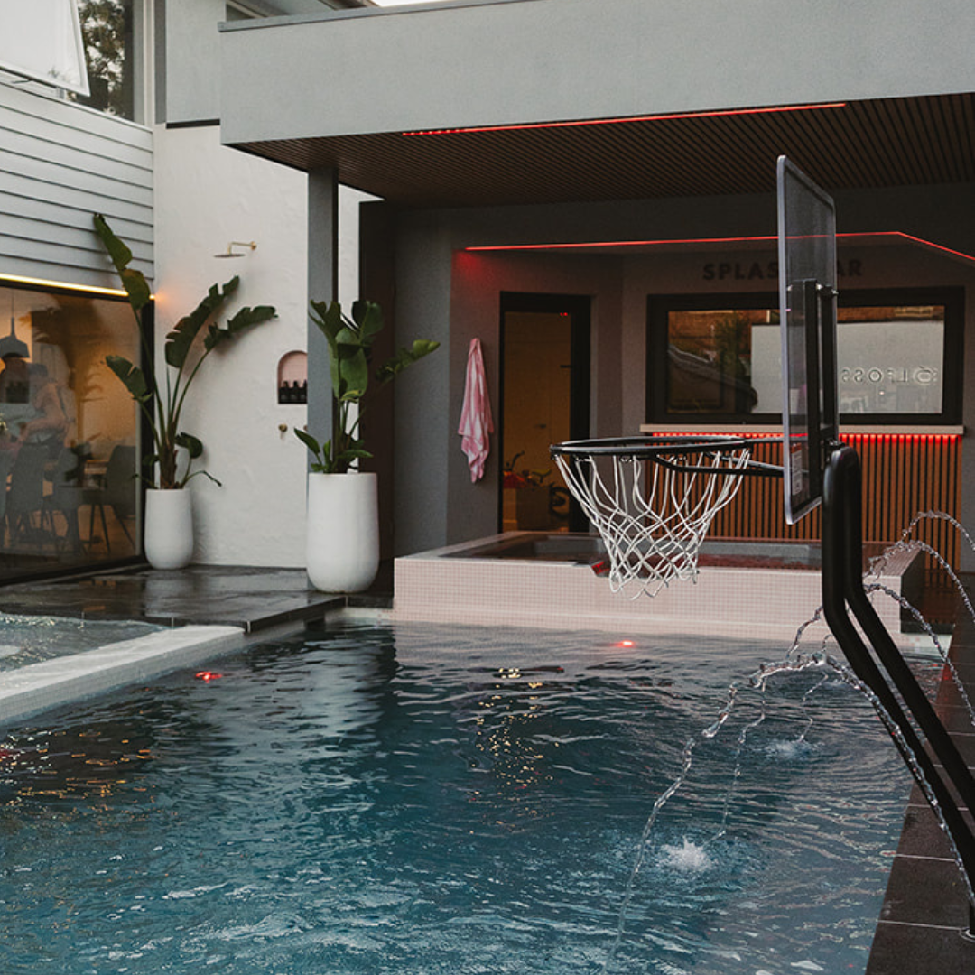 Modern backyard pool featuring an S.R. Smith basketball hoop next to a swim-up bar with LED lighting and integrated water fountains.