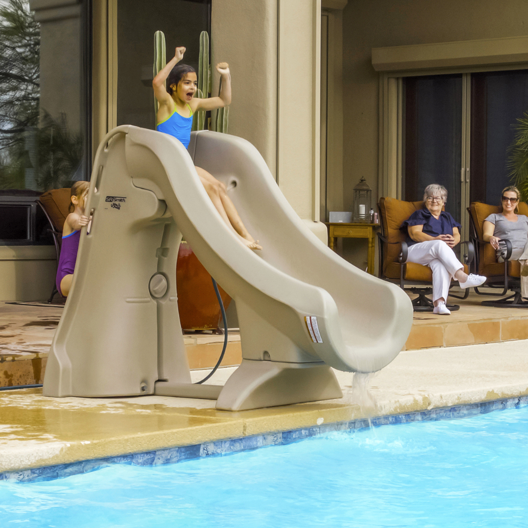 A child slides down a taupe S.R.Smith SlideAway removable inground pool slide while family members watch from patio lounge chairs on a residential deck.