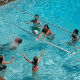 High-angle outdoor lifestyle photo of a family playing water volleyball in a swimming pool with an S.R. Smith Swim N' Spike pool net system across the clear blue water.