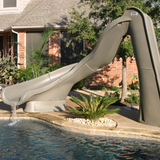 An S.R. Smith TurboTwister gray residential swimming pool slide with an active running water feature flowing into an inground pool, installed on a concrete patio deck with a brick house and landscaped palm plants in the background.