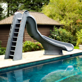 An S.R. Smith TurboTwister gray residential swimming pool slide featuring an integrated deep-rung safety ladder, installed on a flagstone deck of an inground pool with a rustic pool house and green trees in the background.