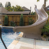 An S.R. Smith TurboTwister sandstone tan residential swimming pool slide with an active water-flow feature pouring into an inground pool, installed on a flagstone patio deck with a wooden privacy fence and residential houses in the background.