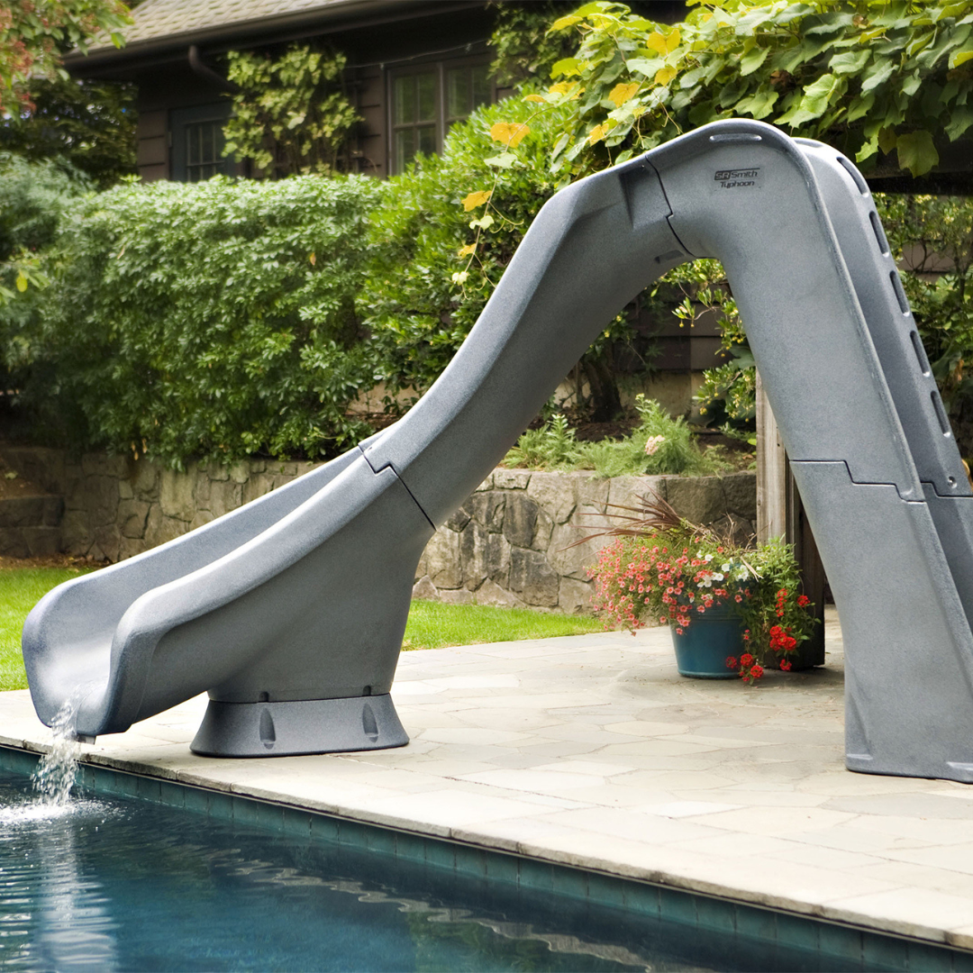 An S.R. Smith Typhoon grey residential swimming pool slide with a running water feature, installed on a flagstone deck of an inground pool in a landscaped backyard with lush green bushes and a stone retaining wall.