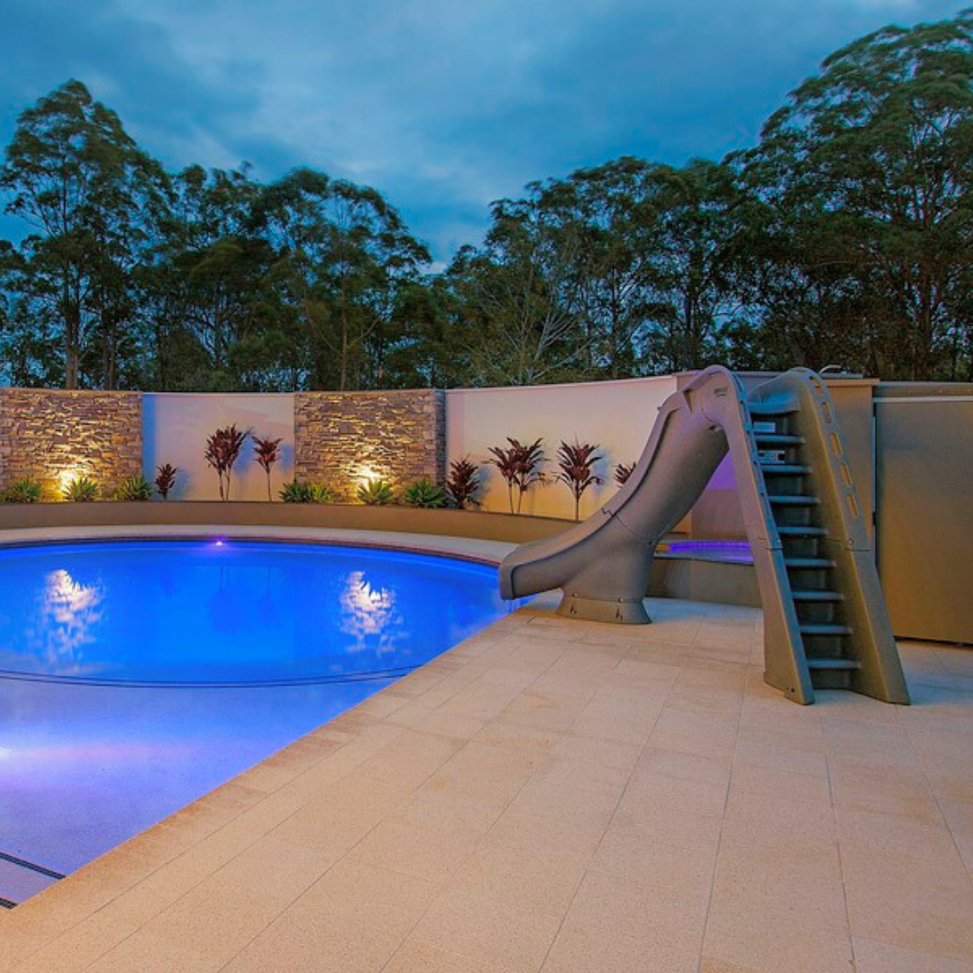 An S.R. Smith Typhoon grey residential swimming pool slide installed on a stone patio next to an illuminated inground pool at twilight, featuring modern stacked stone accent walls, uplighting, and tropical plants in the background.