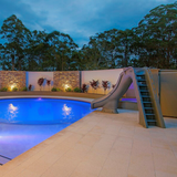 An S.R. Smith Typhoon grey residential swimming pool slide installed on a stone patio next to an illuminated inground pool at twilight, featuring modern stacked stone accent walls, uplighting, and tropical plants in the background.