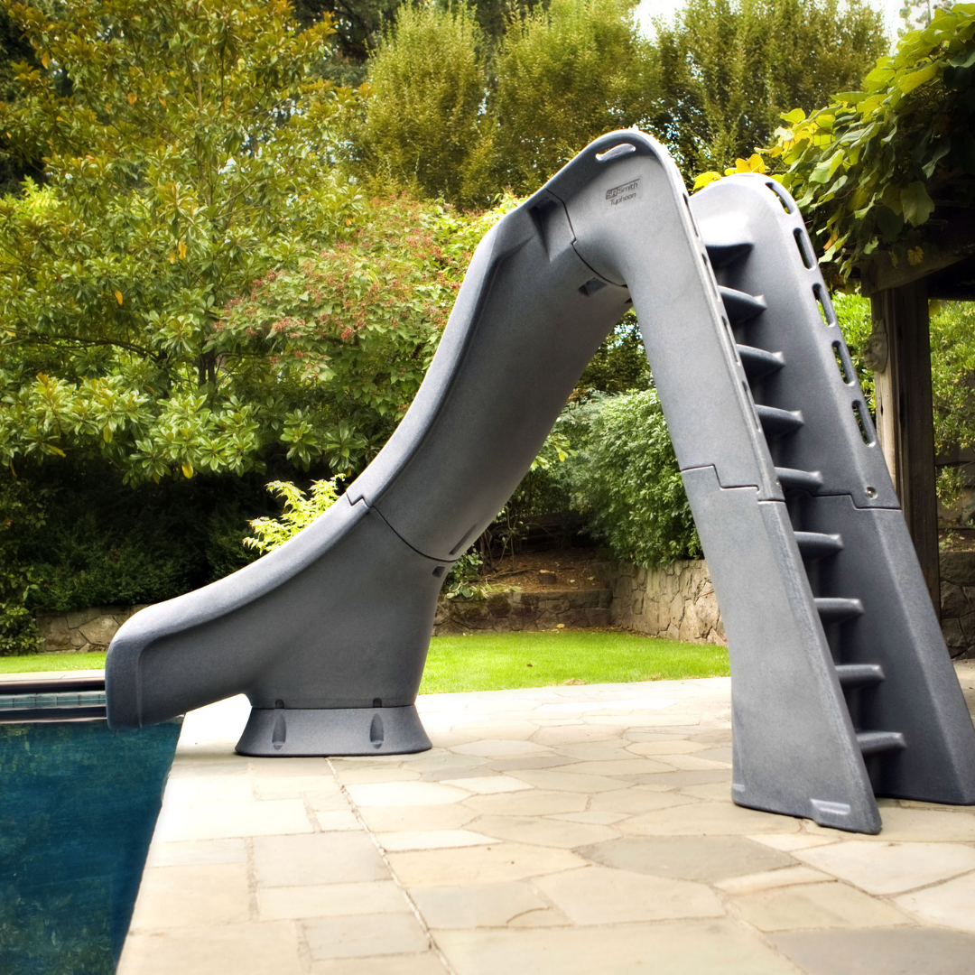 An S.R. Smith Typhoon grey residential swimming pool slide featuring an integrated deep-rung safety ladder, installed on a flagstone pool deck with a lush green backyard and stone wall in the background.