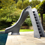 An S.R. Smith Typhoon grey residential swimming pool slide featuring an integrated deep-rung safety ladder, installed on a flagstone pool deck with a lush green backyard and stone wall in the background.