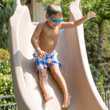 A young boy wearing blue swim goggles and plaid swim trunks sliding down an S.R. Smith Typhoon sandstone tan residential swimming pool slide on a sunny day.