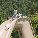 A young boy wearing blue inflatable arm bands sitting at the top of an S.R. Smith Typhoon sandstone tan residential pool slide, getting ready to slide down against a background of lush green trees.