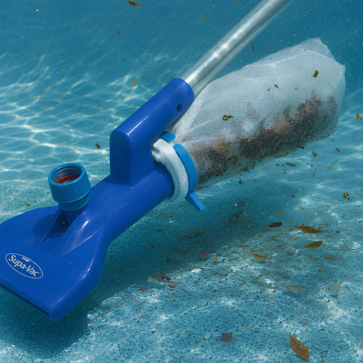 An underwater shot of a blue Supa-Vac pool vacuum being used to clean debris off a textured blue pool floor, showing the attached white mesh filter bag filling up with collected brown leaves and organic waste.