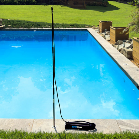 A Swim Tether stationary pool swimming system installed into a deck mount on the stone edge of an outdoor inground backyard swimming pool, showing the vertical black fiberglass pole, resistance cord, and padded swim belt resting on the ground.