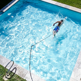 High-angle overhead view of a man swimming freestyle in place using a Swim Tether resistance system attached to a pool deck post in a rectangular backyard swimming pool.