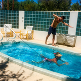 Outdoor lifestyle photo of a woman swimming in place in a small pool using a Swim Tether system attached to a deck pole, while a man on the pool deck gestures and watches, set against a glass block wall backdrop.