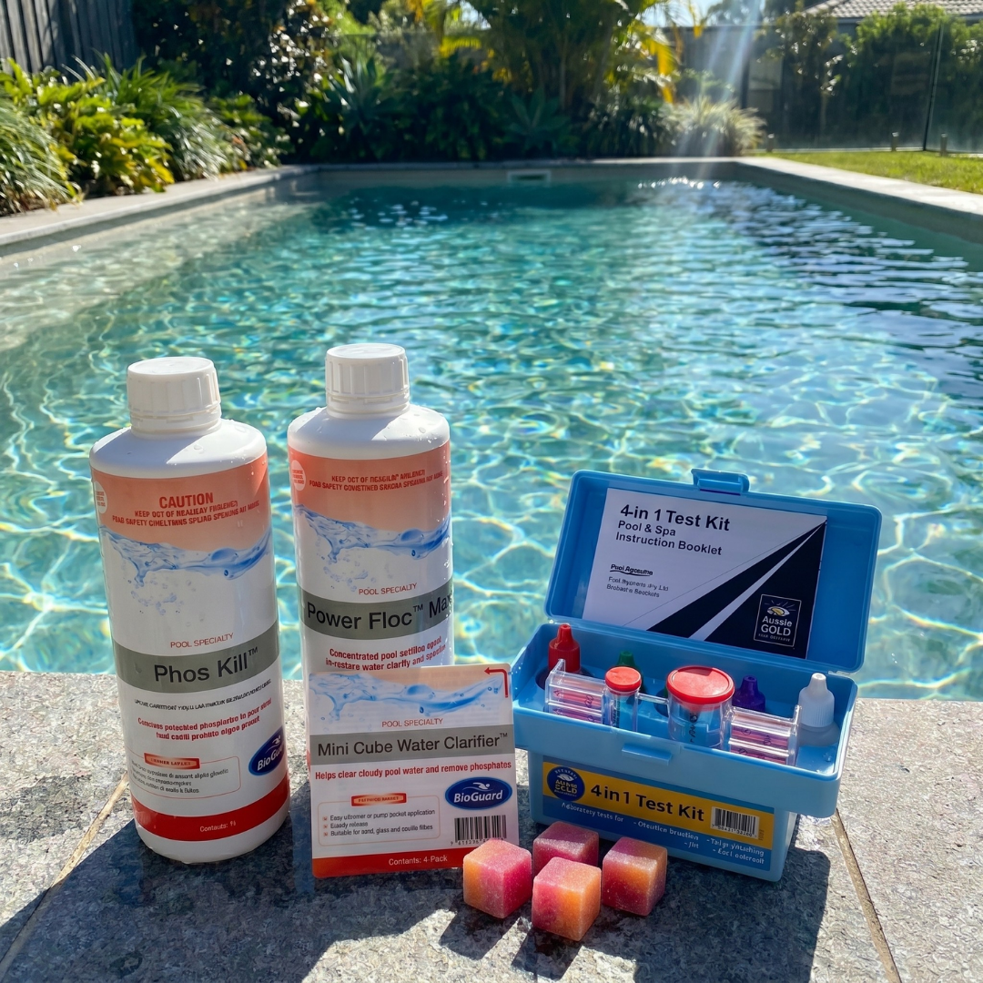 A swimming pool chemical kit on the edge of a clear blue pool, featuring BioGuard Phos Kill, Power Floc Max, Mini Cube Water Clarifier cubes, and an Aussie Gold 4-in-1 Test Kit.