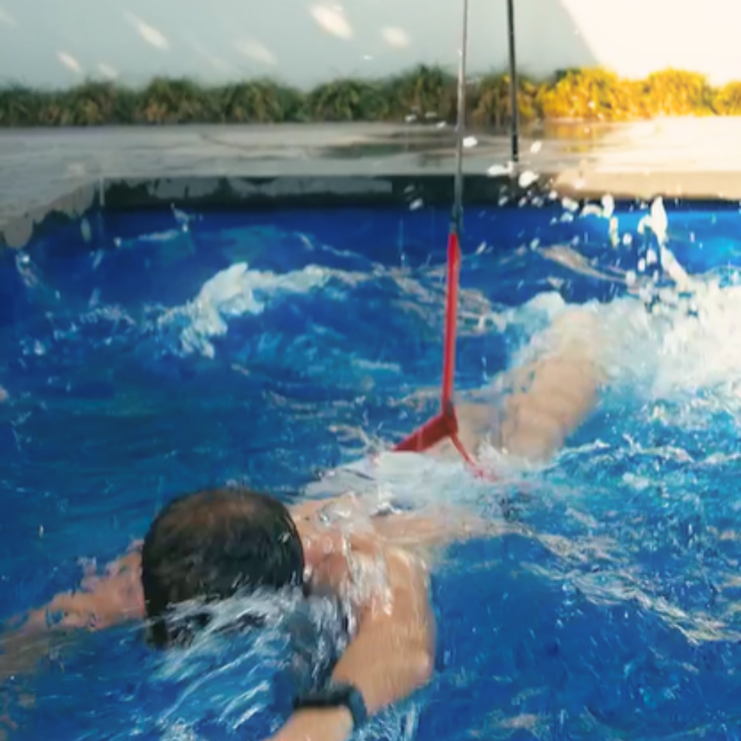 Close-up action shot of an athlete doing a freestyle stroke workout in a swimming pool while attached to a Swim Tether system, showing the red resistance strap and black cord connected to the swimmer's waist.